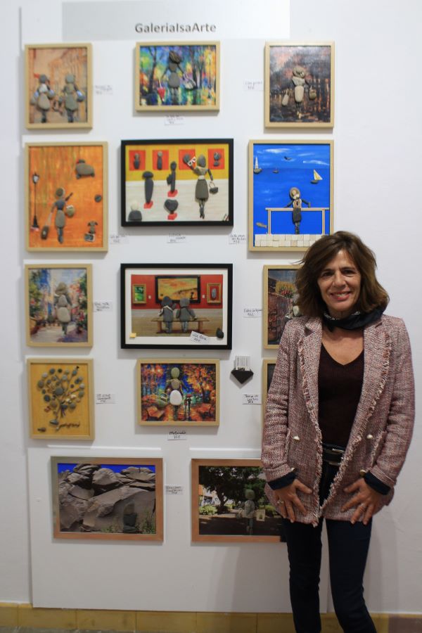 Se ve una fotografía con una mujer sonriente, exponiendo sus cuadros de piedras en una galería, Realmarket21. 