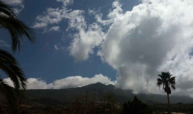 Se ve un paisaje con un fondo de una montaña rodeada de grandes nubes. Se puede apreciar una figura en esas inmensas nubes.