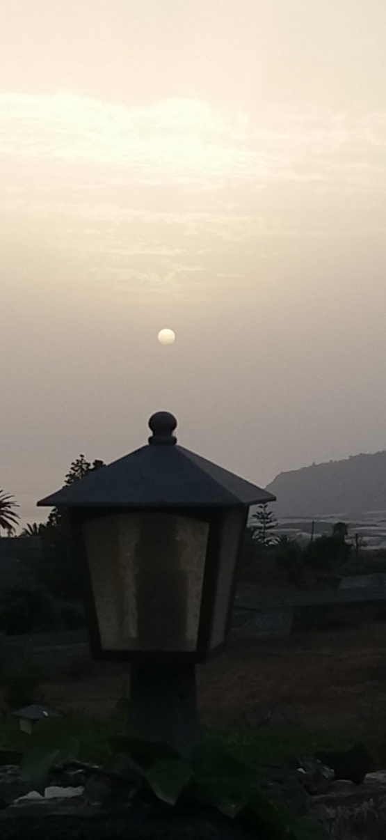 Se ve un atardecer con el sol de calima, encima de un farol, que también ilumina.